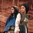 Zwei Frauen in traditioneller Kleidung stehen vor einer felsigen Kulisse, eine trägt ein blaues Tuch, die andere ein reich besticktes Gewand.