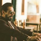 Ein Mann mit Sonnenbrille und Bart sitzt in einem Café. Er trägt einen Anzug und hat konzentrierten Gesichtsausdruck.