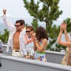 Eine Gruppe gut gekleideter Menschen auf einem Balkon am Meer, klatschend und feiernd. Die Stimmung ist ausgelassen.