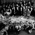Viele Menschen um einen gedeckten Tisch, in der Mitte eine Torte mit Blumen, alle lachen und winken, als Teil eines feierlichen Anlasses.