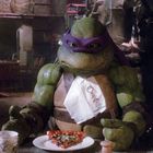 Eine grüne, maskierte Schildkröte sitzt an einem Tisch mit Pizza und Getränk. Sie trägt ein Lätzchen und hält Besteck.