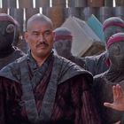 Ein Mann mit kahlrasiertem Kopf in traditioneller Kleidung, umgeben von maskierten Personen mit dunklen Outfits.