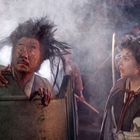 Zwei Personen in einer dichten, nebligen Umgebung. Einer mit wildem Haar schaut überrascht, der andere trägt einen Umhang.