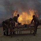 Keine Texte sichtbar. Drei Soldaten tragen ein Bett mit einer Person, während im Hintergrund ein großes Feuer lodert.