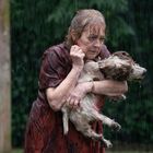 Eine ältere Frau steht im Regen und hält einen nassen Hund fest im Arm. Sie wirkt besorgt, umgeben von unscharfer Vegetation.