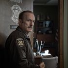 Texte: "SHERIFF," "NORMAL COUNTY," "A. GUNDERSEN." Ein Mann in Uniform steht in einer Tür, hält ein Gebäck.