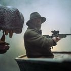 Ein Mann hält ein Gewehr, trägt Hut und Pullover, sitzt in einem Boot. Ein großes, bedrohliches Tiermaul mit Zähnen ist nah bei ihm.