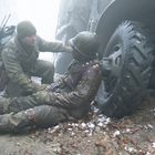 Zwei Soldaten in Winteruniformen; einer kniet und spricht mit dem anderen, der verwundet neben einem Militärfahrzeug liegt.