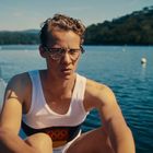 Ein junger Mann in einem Ruderboot trägt ein Sportshirt mit einem olympischen Emblem. Hintergrund: See und Wald.