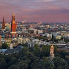 Stadtansicht bei Sonnenuntergang mit einem Turm mit Uhr, vielen Gebäuden und einem Denkmal inmitten von Bäumen.