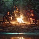 Zwei Personen sitzen am Lagerfeuer im Wald. Im Hintergrund stehen zwei Motorräder.