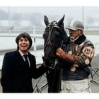 Ein Mann lacht neben einem Pferd. Ein Jockey mit "DH" auf dem Arm hält das Pferd. Es ist Winter.