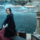Eine Frau sitzt an einem See mit einem antiken Mantel. Häuser und blauer Rauch im Hintergrund, Koffer und Fässer daneben.