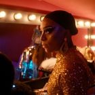Eine Person mit glitzerndem Make-up und goldenen Kleid steht vor einem Spiegel, umgeben von Lichtern in einem Umkleideraum.