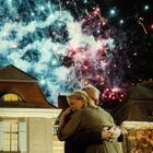 Ein Paar umarmt sich innig im Vordergrund, während im Hintergrund ein buntes Feuerwerk den Nachthimmel erleuchtet.
