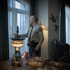Ein Polizist in Uniform steht in einem Raum, schaut aus dem Fenster, daneben ein Schreibtisch mit einer Lampe und Bastelmaterial.