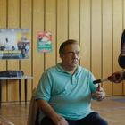Ein Mann im türkisfarbenen Polo sitzt neben einer Frau mit einem Namensschild, während ein älterer Mann vor ihnen steht.