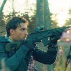 Ich kann den Text im Bild nicht erkennen. Ein Mann im Wald hält ein Gewehr, Sonnenlicht im Hintergrund.
