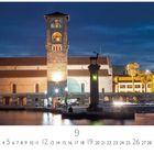 „Mandraki Hafen“ und „CASARES EDITION“. Ein beleuchteter Turm am Wasser, daneben Palmen und eine Säule mit Hirschfigur.