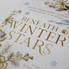 „Beneath Winter Stars“ steht in goldener Schrift, umgeben von floralen Mustern und kleinen blauen Sternen.