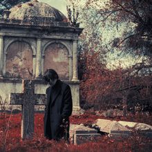 Ein Mann in dunklem Mantel steht nachdenklich vor einem alten Steinkreuz auf einem rot gefärbten Friedhof.