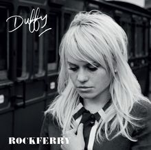 Text: „Duffy“, „ROCKFERRY“. Schwarz-weiß-Foto einer Frau mit blonden Haaren, vor einem Zug stehend, wirkt nachdenklich.