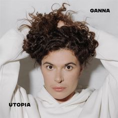 Texte: "GANNA" oben rechts, "UTOPIA" unten links. Person mit lockigem Haar und weißem Hoodie, Hände am Kopf.