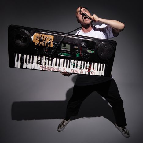Text: "TOMORROW'S WORLD". Eine Person hält ein Keyboard mit elektronischen Komponenten, singt leidenschaftlich ins Mikrofon.