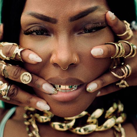 Eine Person mit goldenen Accessoires, Ringen und einem Nasenpiercing. Hände umrahmen Gesicht mit goldenen Ringen.