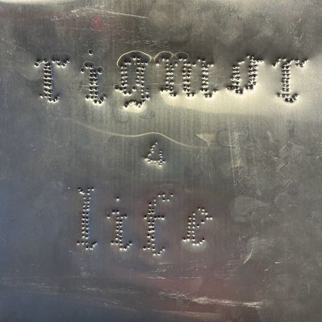 Die Worte "Tigmot 4 life" sind in gepunkteten Löchern auf einer metallischen Oberfläche geprägt.