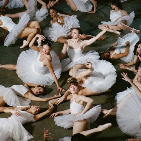 Eine Gruppe von Ballerinas trägt weiße Tutus und posiert anmutig auf dem Boden, teils liegend, teils sitzend.