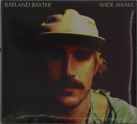 „RAYLAND BAXTER“ und „WIDE AWAKE“ in heller Schrift. Ein Mann mit Schnurrbart und Mütze vor dunklem Hintergrund.