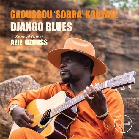 GAOUSSO 'SOBRA' KOUYATE, DJANGO BLUES, Special Guest AZIZ OZOU, ein Mann mit Hut spielt Gitarre vor Felsen.