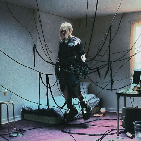 Person in schwarzer Kleidung schwebt mit Kabeln verbunden in einem minimalistisch eingerichteten Zimmer.
