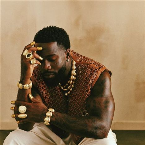Ein Mann sitzt nachdenklich, mit Schmuckringen und -armbändern. Oben rechts Text: "Joyà Tayc 2ème Album. Concerts à l'Accor Arena".