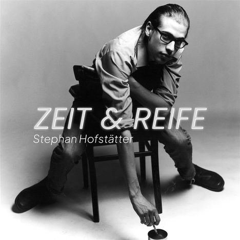 „ZEIT & REIFE“, Stephan Hofstätter. Ein Mann mit Brille sitzt auf einem Stuhl, beugt sich vor und hält eine Zigarette.