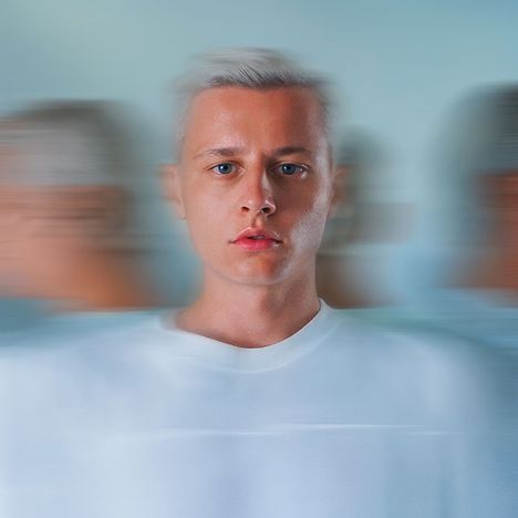Keine Texte erkennbar. Ein Mann mit blondem Haar und weißem Hemd in der Bildmitte, Hintergrund unscharf verwischt.