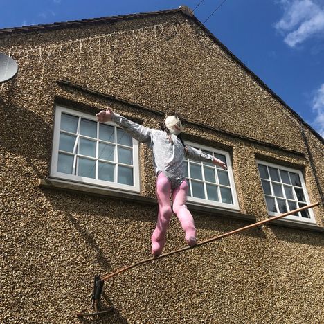 Ein Gebäude mit zwei Fenstern im Obergeschoss. Eine Schaufensterpuppe balanciert auf einem Drahtseil davor.