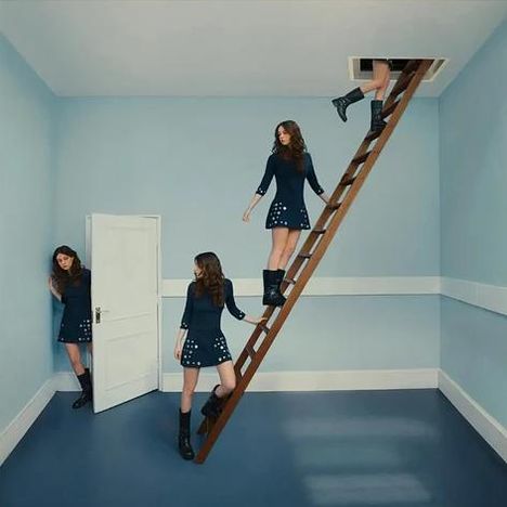 Mehrere Frauen in blauen Kleidern und Stiefeln in einem surrealen Raum mit Tür und Leiter.