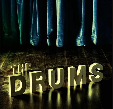 Text: "THE DRUMS". Große 3D-Buchstaben auf einem Holzboden, im Hintergrund ein schwerer, blauer Vorhang.