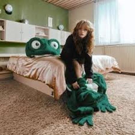 Kein Text sichtbar. 
Eine Person in einem Zimmer mit einem riesigen grünen Frosch-Plüsch auf dem Bett. Gemütliche Atmosphäre.