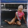 Der Text "FEMME FATALE" steht auf einer Schärpe. Eine Person sitzt vor einem Auto, trägt ein rosa T-Shirt und hat Tattoos.