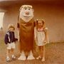 Zwei Kinder posieren draußen mit einer großen Figur in einem braunen Kostüm mit blondem Haar und großer Nase.