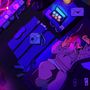 Eine Person liegt in Neonlicht auf einem Bett, hält ein Smartphone, umgeben von Kamera, Socken und Brief mit Herz.