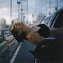 Eine Person lehnt sich lachend aus einem Auto auf einer Brücke. Die Stadt ist im Hintergrund bei Dämmerung zu sehen.