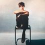 Keine Texte sichtbar. Eine Person sitzt lässig auf einem Stuhl, Arme über den Knien verschränkt. Atmosphäre ruhig.