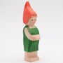 Eine einfache, hölzerne Figur im grünen Outfit mit roter, spitzer Mütze, lächelt freundlich, Arme verschränkt.