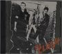 "The Clash" in orange Schrift. Drei Männer in Schwarz-Weiß stehen in einer Gasse. Punk-Ästhetik, abgetragen und roh.