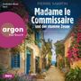 Titel: "Madame le Commissaire und der stumme Zeuge." Die Straße ist gesäumt von Blumen, mit blauen Türen und Fenstern.